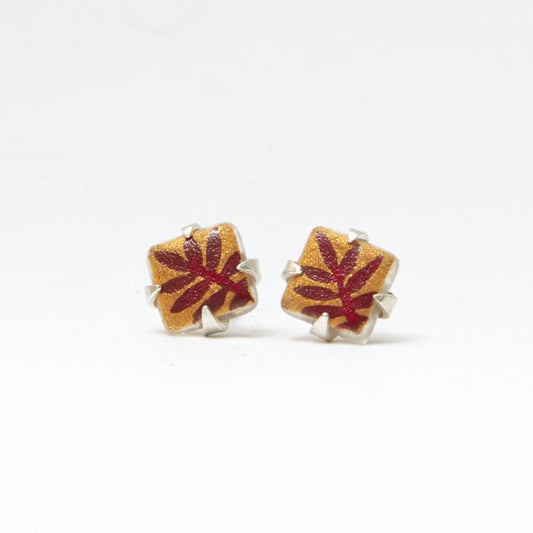 Small square studs crafted with upcycled hand-painted glass, set in sterling silver. Features a botanical pattern for a simple yet elegant design. Golden background and red leaf.