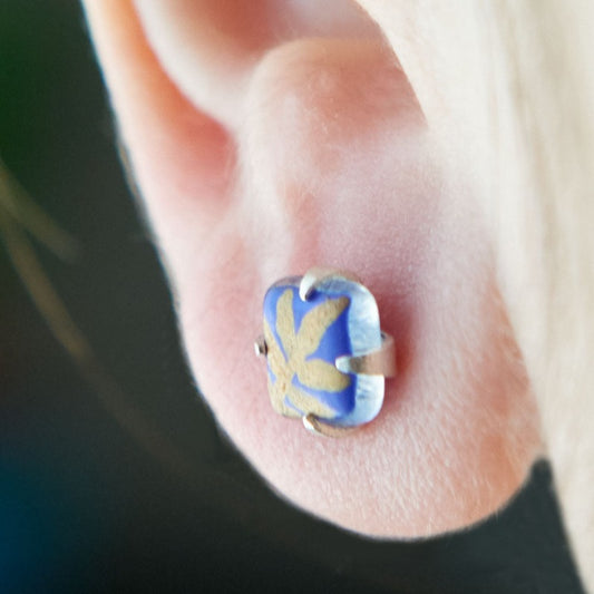 Small square studs crafted with upcycled hand-painted glass, set in sterling silver. Features a botanical pattern for a simple yet elegant design. Blue background and golden leaf.