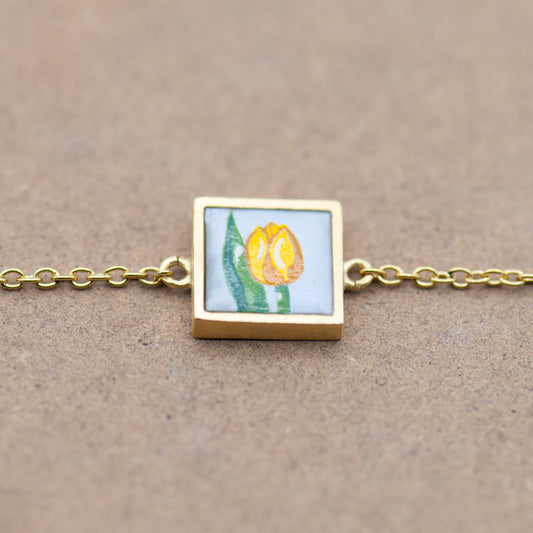 Small golden square frame bracelet, featuring an upcycled hand painted glass of a yellow tulip. Light blue background with a delicate yellow tulip