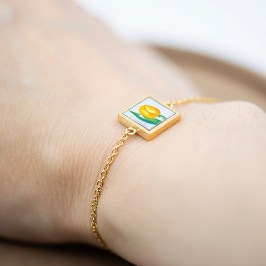Small golden square frame bracelet, featuring an upcycled hand painted glass of a yellow tulip. Light blue background with a delicate yellow tulip