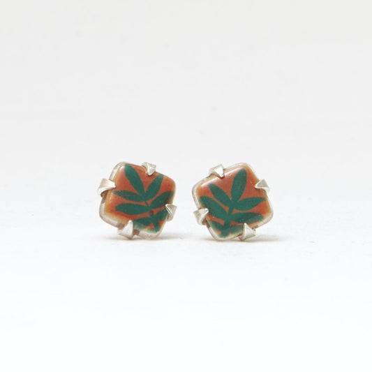 Small square studs crafted with upcycled hand-painted glass, set in sterling silver. Features a botanical pattern for a simple yet elegant design. Orange background and green leaf.