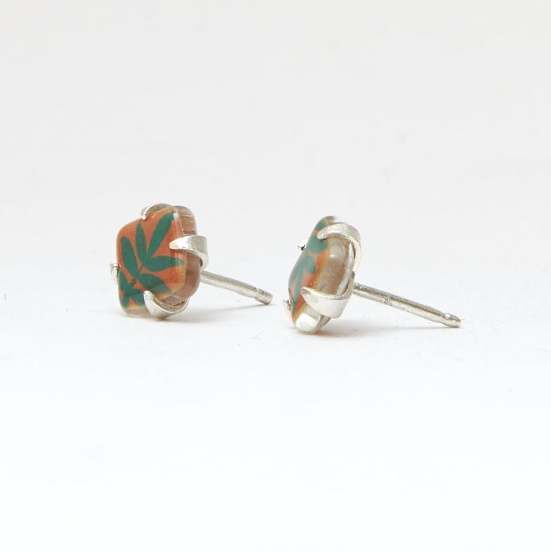 Small square studs crafted with upcycled hand-painted glass, set in sterling silver. Features a botanical pattern for a simple yet elegant design. Orange background and green leaf.