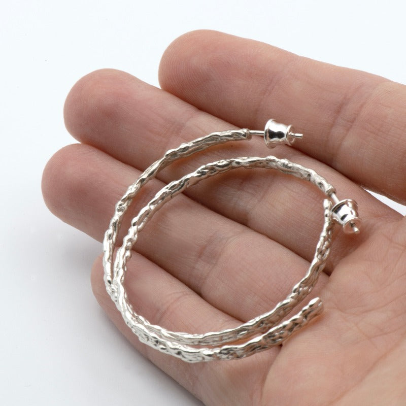 Large round sterling silver hoop earrings with an organic texture reminiscent of sand dripping at the beach. Smooth surface with tactile appeal and visually captivating design.