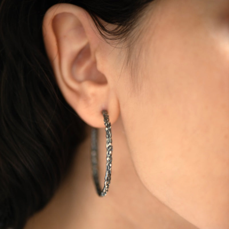 Large round sterling silver hoop earrings with an organic texture reminiscent of sand dripping at the beach. Smooth surface with tactile appeal and visually captivating design. Oxidised silver with contrasting silver and black details for a more sophisticated look.