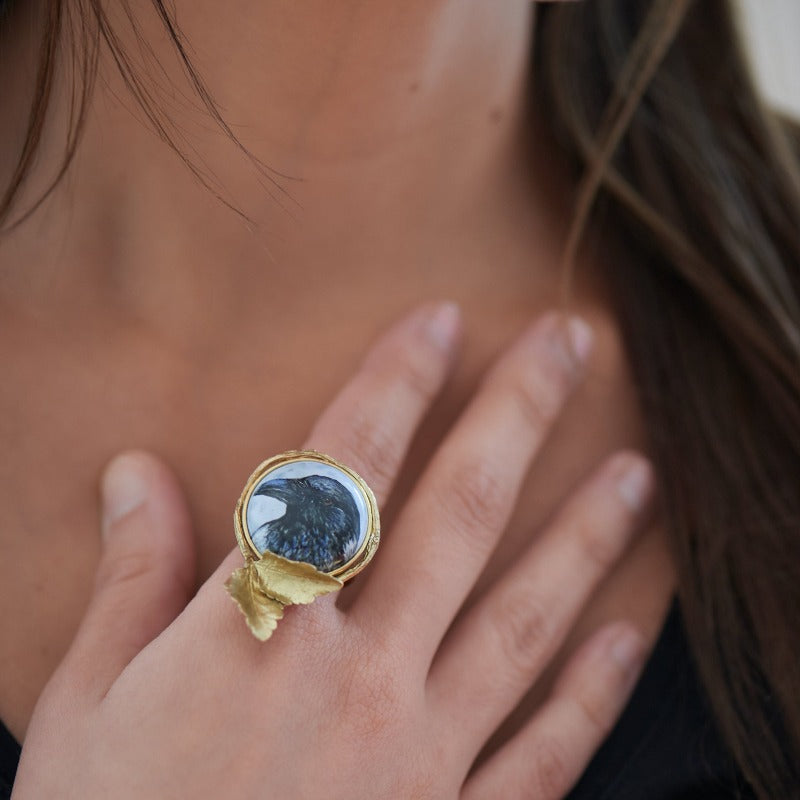 Hand-painted hooded crow head on upcycled glass cabochon, set in an 18k gold-plated, handmade botanical round setting statement ring. Delicate miniature painting setted in a round setting featuring a realistic small tree branch with two small leaves. Light blue background. Realistic adjustable tree branch ring.